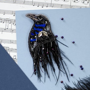 May include: A handcrafted raven brooch, with a black beaded body, blue accents, and a silver beak. The bird design includes black feathers and small purple beads, set against a light blue background with musical notes.