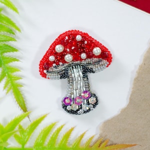 May include: A beaded mushroom brooch with a red cap dotted with white pearls and beads. The stem is silver and gray, with a black base adorned with sequins. The brooch is a whimsical accessory.