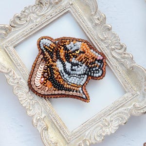May include: A beaded tiger brooch in shades of orange, black, and white, set within a white ornate frame. The brooch is surrounded by other frames and scattered sequins. The overall theme is handcrafted jewelry.