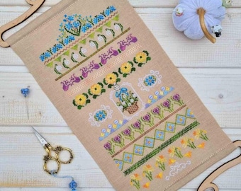 May include: A beige cross-stitch sampler with floral patterns in blue, purple, yellow, and green, displayed on a wooden scroll frame. A white pincushion with pins, a blue tassel, and gold scissors are nearby.