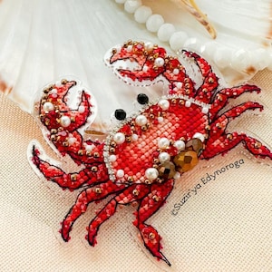 May include: A red crab brooch with intricate beadwork and pearl accents. The crab's body is a vibrant red, with detailed legs and claws. The brooch is set against a seashell and a neutral background.