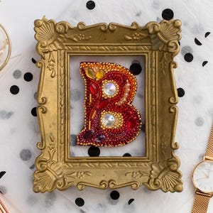 May include: A gold-framed, beaded letter "B" in red and orange, with clear crystal accents. A gold watch, perfume, earrings, and pencils are arranged on a white surface with black polka dots and a card that says "You are beautiful."