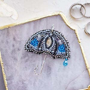 May include: A beaded umbrella brooch with silver, blue, and gray beads, set on a gold-edged, marble-like tray. A blue teardrop bead hangs from the umbrella. Rings and a candle are in the background.