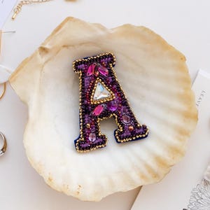 May include: A beaded letter "A" brooch in shades of purple, pink, and gold, resting in a seashell. The brooch is surrounded by gold jewelry, including hoop earrings, rings, and a necklace, on a white surface.