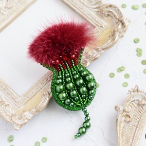 Puede incluir: Broche artesanal con forma de cardo. Presenta una parte superior de plumas burdeos, un cuerpo verde perlado y un tallo perlado. El broche está sobre un fondo blanco con marcos decorativos y lentejuelas verdes.