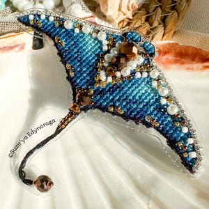May include: A handcrafted, denim-textured stingray brooch. The brooch features intricate beadwork in shades of blue, brown, gold, and white, with a clear plastic backing. The design includes a beaded tail and is embellished with rhinestones and pearls.