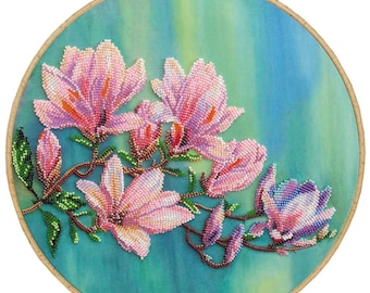 May include: A circular embroidery of pink magnolia flowers with orange centres and green leaves, set against a teal and blue backdrop. The wooden frame has a brass screw. The artwork is approximately 25 cm in diameter.