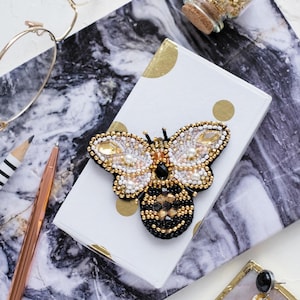May include: A beaded bee brooch with black, white and gold accents. The bee is sitting on a white and gold notebook with black polka dots.