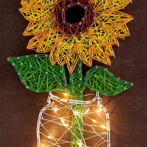 Könnte beinhalten: Handgefertigte String-Art-Arbeit, die eine Sonnenblume in einem Einmachglas darstellt. Die Sonnenblume hat gelbe und orangefarbene Fäden mit einem braunen Zentrum. Grüne Fäden bilden die Blätter und den Stiel. Das Glas wird von weißen Lichterketten beleuchtet.