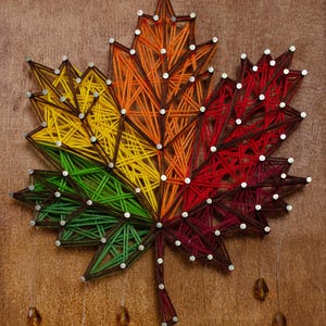 Puede incluir: Una colorida pieza de arte de hilo de hoja de arce. La hoja está dividida en secciones de hilo verde, amarillo, naranja y rojo, aseguradas con pequeños clavos sobre un fondo de madera. Cuentas decorativas cuelgan debajo.