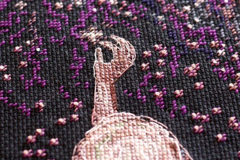 Pu&ograve; includere: Primo piano di un disegno a punto croce con un gesto della mano con l'indice rivolto verso l'alto. La mano &egrave; cucita in tonalit&agrave; marroni e beige, su uno sfondo nero con accenti floreali viola.