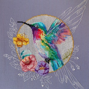 May include: A colorful cross-stitch embroidery of a hummingbird with iridescent green and blue feathers, a pink and red chest, and a long, thin beak. The hummingbird is surrounded by a wreath of white flowers and leaves, and a gold circle.
