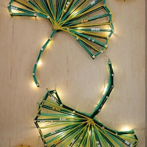 Peut inclure: Art de ficelle représentant des feuilles de ginkgo avec des fils jaunes, verts et noirs sur un fond en bois. L'œuvre comprend de petites perles blanches et dorées et de petites lumières. Deux petites feuilles de ginkgo sont également présentes.