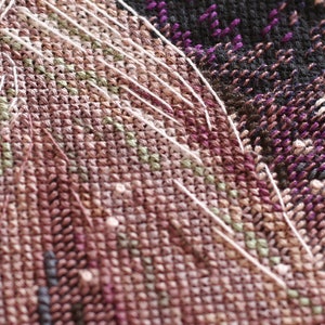 Pu&ograve; includere: Primo piano di un tessuto a punto croce con un motivo complesso. Il tessuto presenta una miscela di colori, tra cui tonalit&agrave; di marrone, viola, verde e bianco. Fili bianchi si estendono sulla superficie, aggiungendo consistenza al design. La cucitura &egrave; fitta e dettagliata.