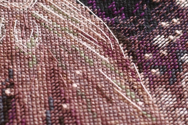 Pu&ograve; includere: Primo piano di un tessuto a punto croce con un motivo complesso. Il tessuto presenta una miscela di colori, tra cui tonalit&agrave; di marrone, viola, verde e bianco. Fili bianchi si estendono sulla superficie, aggiungendo consistenza al design. La cucitura &egrave; fitta e dettagliata.