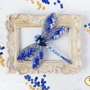 May include: A blue and gold dragonfly brooch made of beads and sequins, displayed in a decorative frame. The brooch is surrounded by blue and gold sequins.