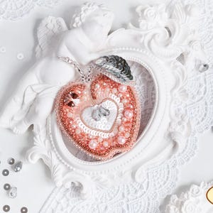 Op de afbeelding: Een delicaat roze en wit parelhartvormig ornament met een zilveren accent. Het ornament wordt getoond in een wit, versierd frame.