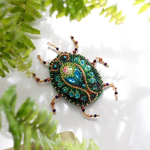 May include: A green and blue jeweled beetle brooch with gold accents. The brooch is made of beads and rhinestones.