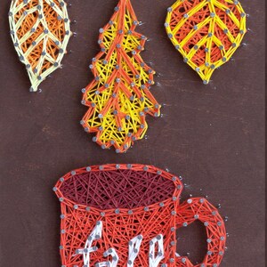 Puede incluir: Arte de hilo que presenta tres hojas de otoño en amarillo, naranja y crema, y una taza roja con la palabra "fall" en blanco. La obra de arte está hecha con clavos e hilo sobre un fondo de madera marrón.