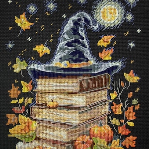 May include: A witch's hat sits atop a stack of books with pumpkins and fall leaves scattered around the base. The hat is blue and gray with a gold band. The books are brown and have gold lettering. The pumpkins are orange and the leaves are red, orange, and yellow. The background is black with a full moon and stars.