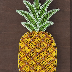 Op de afbeelding: Een ananas van draadkunst op een donkerbruine houten achtergrond. De ananas heeft een gele body met bruine accenten en groene en witte bladeren. Het ontwerp is gemaakt met draad en kleine zilveren spijkers.