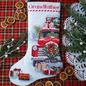 Può includere: Una calza natalizia bianca con un disegno a punto croce di un camion rosso vintage che trasporta regali incartati. Il camion ha una ghirlanda verde sulla griglia anteriore. La parola Grandfather è ricamata in rosso sulla parte superiore.