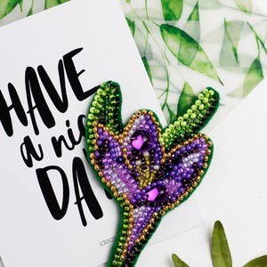 Könnte beinhalten: Handgefertigte Blumenbrosche mit Perlen in Lila, Grün und Gold. Die Brosche ist mit kleinen Perlen und Edelsteinen verziert. Eine weiße Karte mit dem Text "HAVE a nice DAY" befindet sich im Hintergrund.