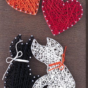 以下が含まれることがあります： 木製の壁掛けアートで、ストリングアートのハートと猫のシルエットが特徴です。2つのハートはオレンジと赤の糸でできており、2匹の猫は黒と白の糸でできています。猫は互いに向き合い、首にリボンを結んでいます。