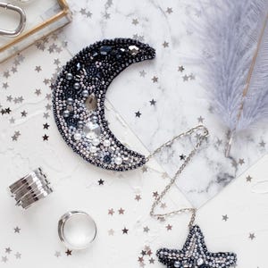 May include: A silver chain necklace with a beaded crescent moon and star pendant. The moon and star are black and silver and have a sparkly, beaded design. The necklace is on a white marble surface with silver glitter.