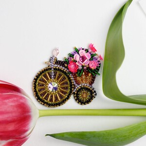 May include: A beaded bicycle brooch with a basket of pink and purple flowers. The bicycle wheels are gold and black, with a large crystal in the center. The brooch is set against a white background with a red tulip and green leaf.