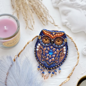 May include: A beaded owl brooch with blue, gold, and orange accents. The owl has a large blue gemstone in its chest and a beaded fringe hanging from its bottom.