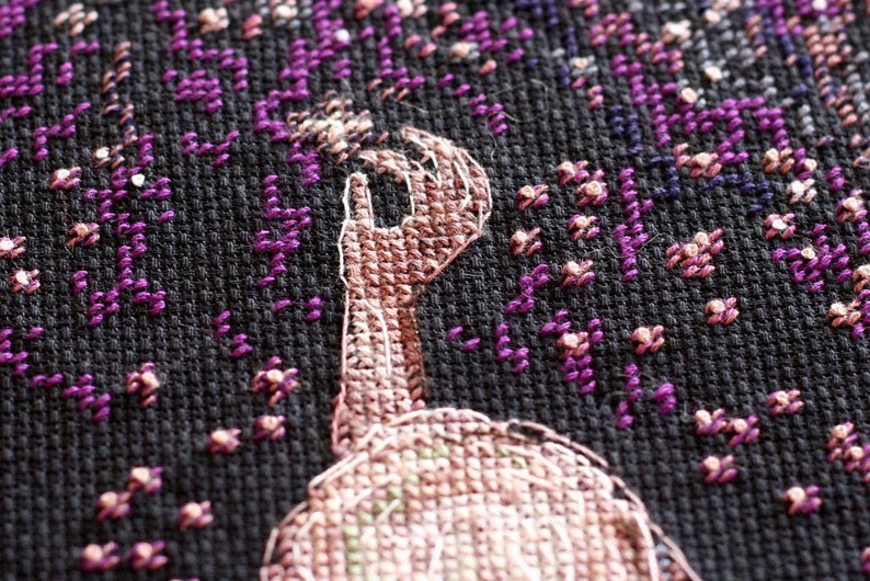 Pu&ograve; includere: Primo piano di un disegno a punto croce con una mano con l'indice rivolto verso l'alto. La mano &egrave; cucita in tonalit&agrave; marroni e beige, su uno sfondo nero con accenti floreali viola e rosa. Il ricamo &egrave; dettagliato.