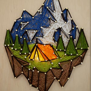 May include: A string art piece depicting a camping scene. The artwork features a mountain range in blue and white, green trees, and an orange tent. The piece is made with string and nails on a wooden background.