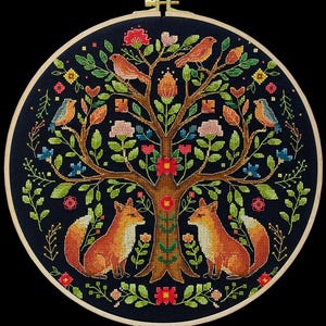 May include: Cross-stitch embroidery in a wooden hoop. The design features a tree with birds, flowers, and two foxes. The color palette includes shades of orange, green, blue, and pink against a dark blue background. The hoop is approximately 25 cm in diameter.