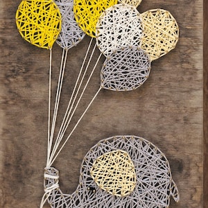 May include: String art elephant with balloons in yellow, gray, and white on a wooden background. The elephant and balloons are made with string and nails, creating a unique, handmade piece of art.