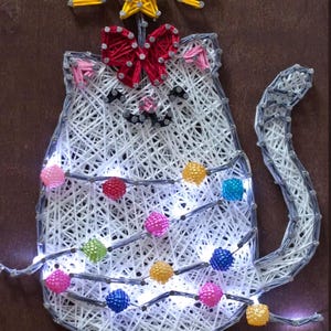 May include: A string art cat silhouette with a red bow, star, and colorful lights. The cat is made of white string on a brown background. The lights are multi-colored and strung across the cat's body.