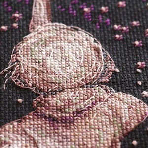 Pu&ograve; includere: Primo piano di un disegno a punto croce raffigurante una figura con cappello e colletto, resa in tonalit&agrave; marroni, rosa e verdi. Lo sfondo nero presenta piccoli accenti floreali viola e bianchi sparsi, creando un'estetica artigianale strutturata.