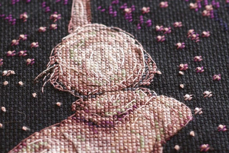Pu&ograve; includere: Primo piano di un disegno a punto croce raffigurante una figura con cappello e colletto, resa in tonalit&agrave; marroni, rosa e verdi. Lo sfondo nero presenta piccoli accenti floreali viola e bianchi sparsi, creando un'estetica artigianale strutturata.