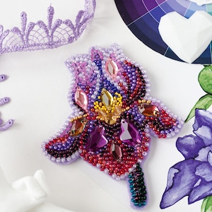 May include: A beaded flower brooch in shades of purple, pink, red, and yellow. The flower is made with small beads and rhinestones, and it has a green stem.
