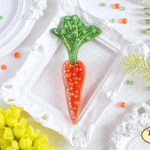 May include: A beaded carrot brooch with green leaves, displayed in a white frame on a white background with green and orange beads scattered around it.