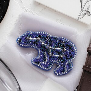 May include: A blue beaded polar bear brooch with a constellation design. The brooch is made with various shades of blue and silver beads, and is set on a white fabric. The brooch is a unique accessory.