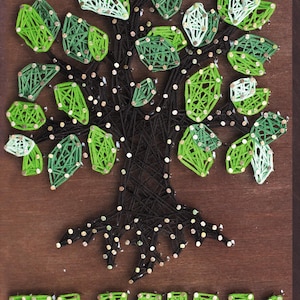 Peut inclure: Une œuvre d'art murale en bois représentant un arbre réalisé avec du fil noir et de la laine verte. L'arbre a des feuilles vertes et le mot "FAMILY" est épelé en laine verte en bas. Le fond est marron foncé.