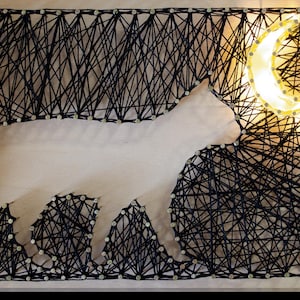Puede incluir: Una pieza de arte de hilo hecha a mano que presenta la silueta de un gato blanco sobre un fondo de hilo negro. Una luna creciente brillante está en la esquina superior derecha. La obra de arte está montada en un marco de madera.