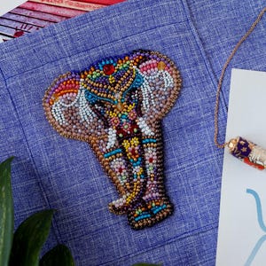 May include: A beaded elephant brooch in shades of brown, blue, red, and yellow, resting on a blue linen surface. The brooch features intricate beadwork and a red gem. A gold chain with a decorative pendant is also visible.