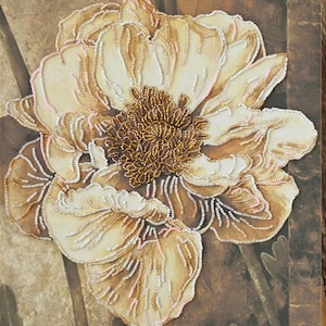 May include: A close-up of a beige and brown floral artwork. The flower has multiple layers of petals, with a center of small, golden beads. The petals are outlined with white and gold beads, and the background is a textured brown.
