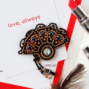 Könnte beinhalten: Eine fächerförmige Brosche mit schwarzen, goldenen und juwelenfarbenen Perlen. Die Brosche hat einen zentralen irisierenden Stein und eine Perlenquaste. Im Hintergrund steht der Text "love, always".