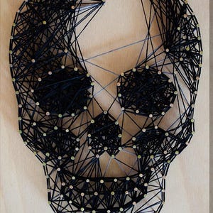 May include: A string art skull design on a light wood background. The skull is created with black thread strung between small nails, forming the shape of a skull. The design is a unique piece of wall decor.