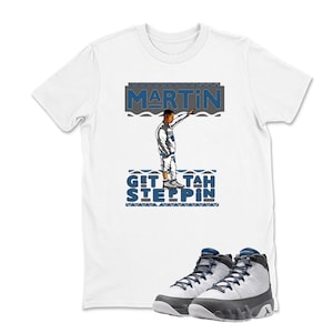May include: White t-shirt with a graphic of a person and the text "MARTIN" and "GIT TAH STEPPIN" in blue and gray. A pair of white, gray, and blue sneakers are in the foreground.