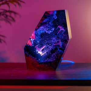 May include: A unique, geometric-shaped lamp with a space-themed design. The lamp features a dark, galaxy-like interior with miniature starfighters. The exterior is a mix of red, blue, and black resin. A blue power cord is visible.