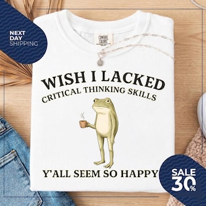 May include: A white t-shirt featuring a graphic of a standing green frog holding a small brown mug. The text above the frog reads WISH I LACKED CRITICAL THINKING SKILLS and the text below the frog reads Y'ALL SEEM SO HAPPY in black lettering.
